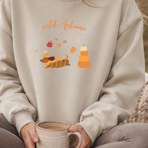 Könnte beinhalten: Ein beigefarbenes Sweatshirt mit Rundhalsausschnitt und dem Schriftzug "wild Autumn" in orangefarbener Schreibschrift. Das Motiv zeigt einen braunen Dackel mit gelber Mütze und Schal, umgeben von Herbstblättern und drei gestapelten Kürbissen.