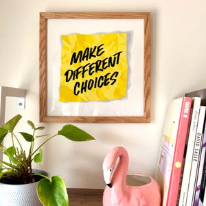 May include: A framed print of a yellow sticky note with the text "MAKE DIFFERENT CHOICES" written in black. The sticky note is crumpled and has a shadow behind it.