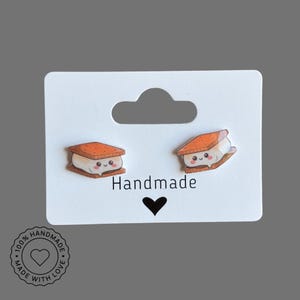Könnte beinhalten: Ein Paar handgefertigte S'mores-Ohrringe auf einer weißen Karte. Jeder Ohrring zeigt ein lächelndes S'more mit einem Graham-Cracker-Ober- und Unterteil und einer Marshmallow-Mitte. Auf der Karte steht das Wort "Handmade" und ein Herzsymbol.
