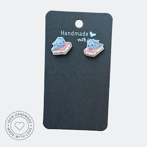 May include: A pair of handmade stud earrings featuring a blue dragon resting on a pink book. The earrings are displayed on a black card with the text "Handmade with love". A circular sticker reads "100% Handmade with Love".