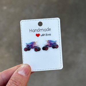 May include: A pair of handmade unicorn monster truck earrings on a white card. The earrings are purple, blue, and pink with red wheels and gold horns. The card reads "Handmade with love" in black text.