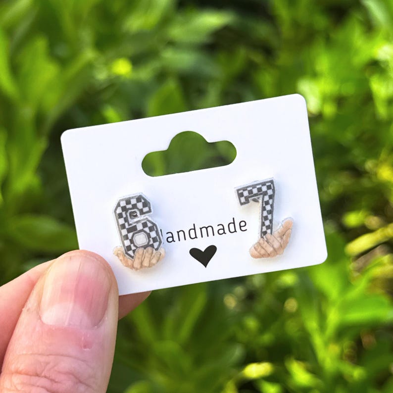 May include: Earrings featuring the numbers 6 and 7 in a checkered pattern, with a small hand design. The earrings are displayed on a white card with the word "handmade" and a black heart.