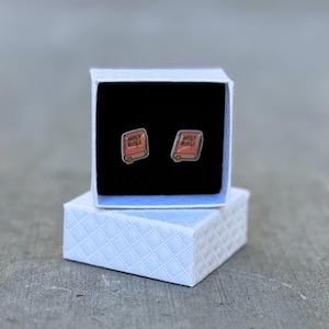 May include: A pair of stud earrings shaped like miniature Bibles, each with the words "Holy Bible" on the cover. The earrings are displayed in an open, white, square gift box with a black interior. The box sits atop a matching, closed box.