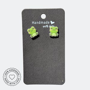 May include: A pair of handmade frog earrings on a black display card. The earrings are green with smiling faces and are labeled "Handmade with love". The card also has a circular stamp that says "100% Handmade with love".