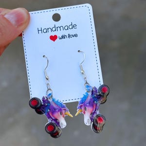 May include: A pair of unicorn earrings with a monster truck design. The earrings are purple, blue, and white with red wheels. The card behind the earrings says "Handmade with love".