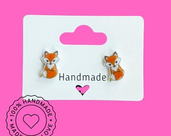 May include: A pair of fox-shaped earrings on a white card with the word "Handmade" and a heart. The earrings are orange and white with green accents. A circular sticker reads "100% Handmade with Love."