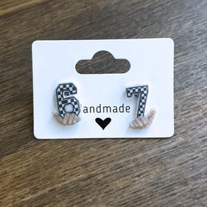 May include: A pair of earrings featuring the numbers 6 and 7 in a black and white checkered pattern, held by small hand illustrations. The earrings are displayed on a white card with the word "handmade" and a black heart.