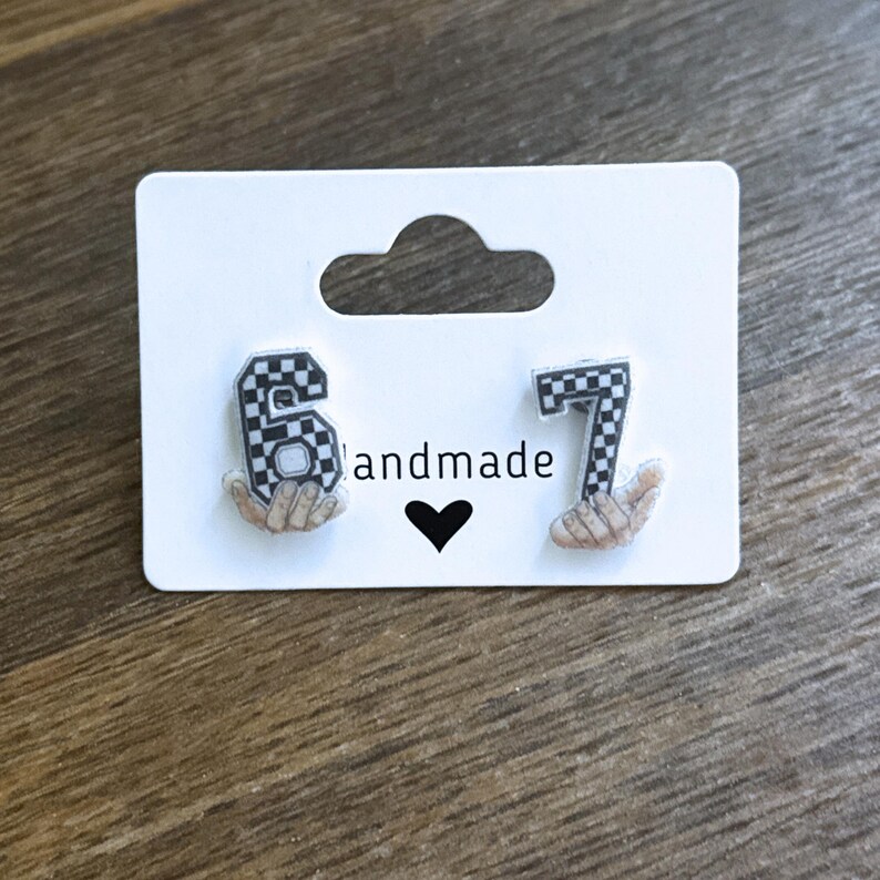 May include: A pair of earrings featuring the numbers 6 and 7 in a black and white checkered pattern, held by small hand illustrations. The earrings are displayed on a white card with the word "handmade" and a black heart.