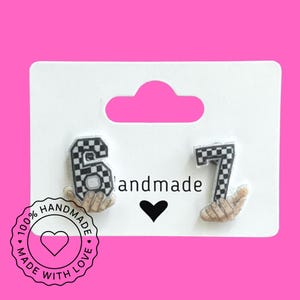 May include: Earrings featuring the numbers 6 and 7 in a black and white checkered pattern, held by hands. The card reads "handmade" with a heart symbol. A circular sticker says "100% handmade with love."