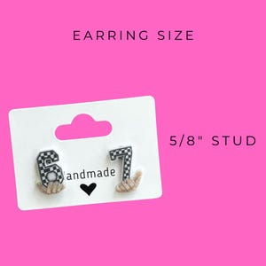 May include: A pair of stud earrings featuring the numbers 6 and 7 in a black and white checkered pattern. The earrings are 5/8" studs and are displayed on a white card with the word "handmade" and a heart symbol. The background is bright pink.