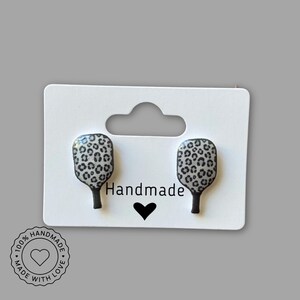 May include: A pair of handmade earrings shaped like pickleball paddles. The paddle faces have a black and white leopard print design. The earrings are displayed on a white card with the word "Handmade" and a heart symbol.
