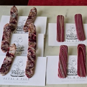 May include: An assortment of handcrafted, elongated stone pieces in shades of red, brown, and cream, arranged on white cards. The cards feature the text "PETAL & PAGE" and "HANDMADE CRAFTS AND DESIGNS".