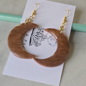 May include: A pair of crescent moon earrings. The earrings are a marbled brown color with gold flecks and gold-colored ear wires. The earrings are hanging on a white card with a floral design.