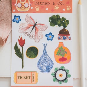 May include: A sheet of stickers featuring a cat with glasses, a butterfly, a potted plant, a vase, a flower, a ticket, and a flower with a green center. The stickers are on a pink background with white paw prints and the text "Catnap & Co."