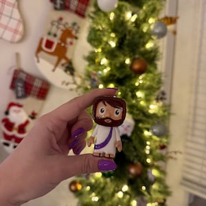 May include: A small, hand-painted figurine of a religious figure, featuring a white robe, brown hair and beard, and a purple sash. The figurine is held in front of a decorated Christmas tree with ornaments and lights. Christmas stockings are visible in the background.