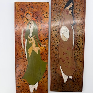 May include: Two rectangular wall plaques featuring painted figures on a mottled brown and black background. The left plaque shows a figure in a green dress and orange headscarf. The right plaque depicts a figure in a brown floral dress.