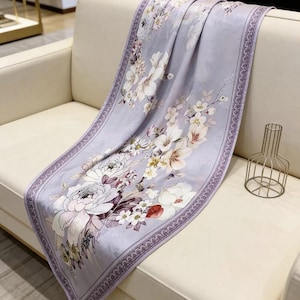 Puede incluir: Una bufanda lavanda con un estampado floral de flores blancas, rosas y rojas. La bufanda tiene un borde decorativo y está drapeada sobre un sofá blanco. La tela es suave y fluida.