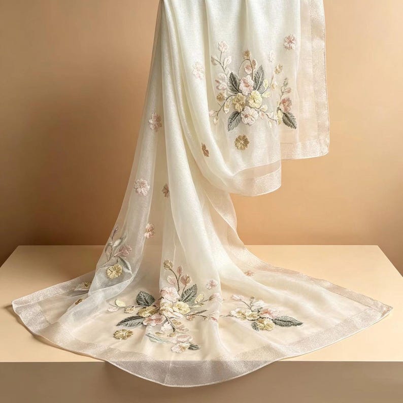 May include: Ivory-colored scarf with floral embroidery. The scarf features detailed embroidery of flowers in shades of pink, yellow, and green. The scarf is made of a sheer, lightweight fabric, and is draped over a surface.
