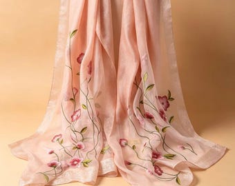 May include: A sheer, light peach-colored scarf featuring delicate floral embroidery. The design showcases climbing vines with small green leaves and pink blossoms scattered across the fabric. The scarf has a subtle, shimmering texture.