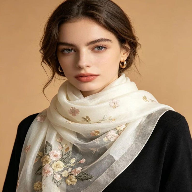 May include: A cream-colored scarf with floral embroidery. The scarf features delicate pink, yellow, and green flower designs. The scarf is draped over a black top.