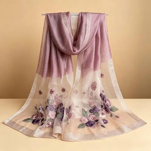 May include: A sheer, mauve-colored scarf with embroidered floral details. The scarf features a gradient design, transitioning from a solid mauve to a sheer, off-white fabric. The edges are trimmed with a silver metallic border, and the scarf is draped over a metal rod.