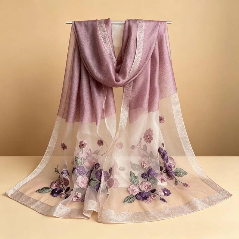 May include: A sheer, mauve-colored scarf with embroidered floral details. The scarf features a gradient design, transitioning from a solid mauve to a sheer, off-white fabric. The edges are trimmed with a silver metallic border, and the scarf is draped over a metal rod.