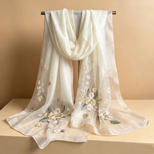 May include: A sheer, ivory-colored scarf with delicate floral embroidery in soft pink, yellow, and green hues. The scarf is draped over a metal bar, showcasing its flowing fabric and intricate design, perfect for adding a touch of elegance.
