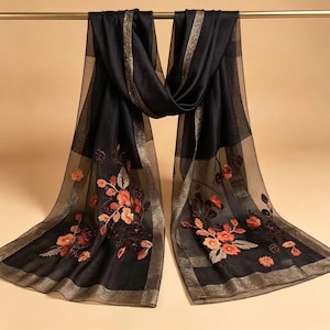 May include: Black scarf with floral embroidery in shades of orange and red. The scarf has a sheer, lightweight appearance with gold trim along the edges and a gold stripe down the center. The scarf is hanging from a gold bar.