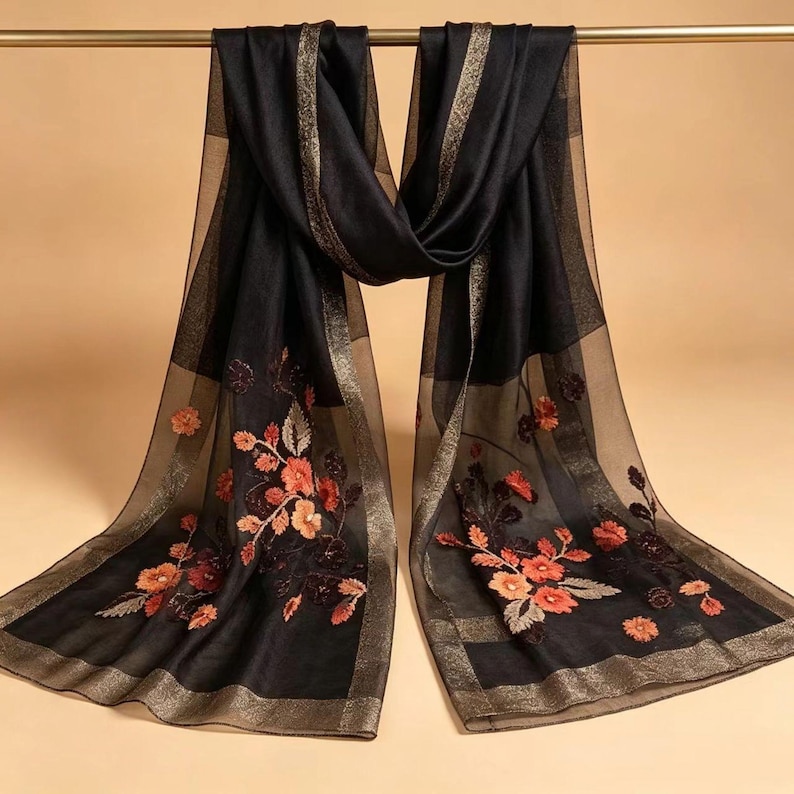 May include: Black scarf with floral embroidery in shades of orange and red. The scarf has a sheer, lightweight appearance with gold trim along the edges and a gold stripe down the center. The scarf is hanging from a gold bar.