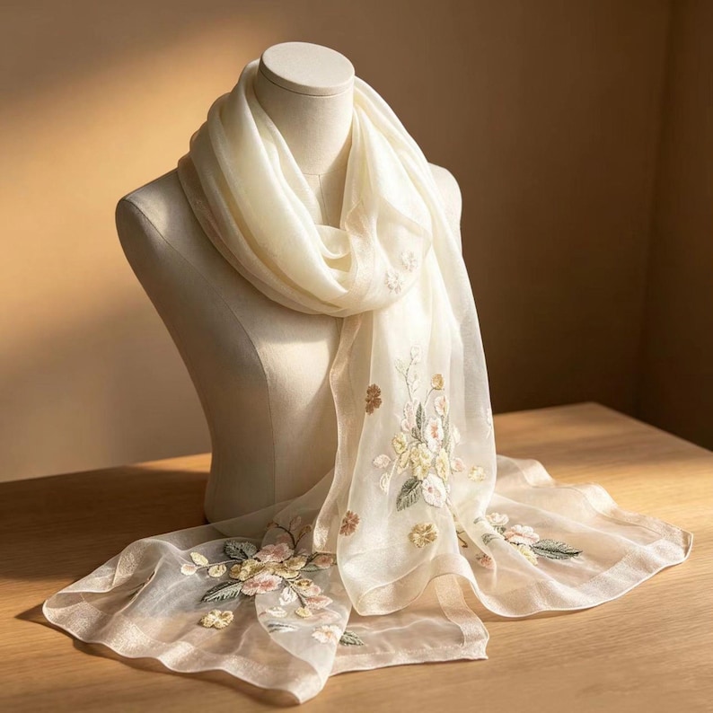 May include: A cream-colored scarf with embroidered floral details. The scarf is draped over a mannequin, showcasing its delicate, sheer fabric and intricate flower patterns in shades of pink, yellow, and green.