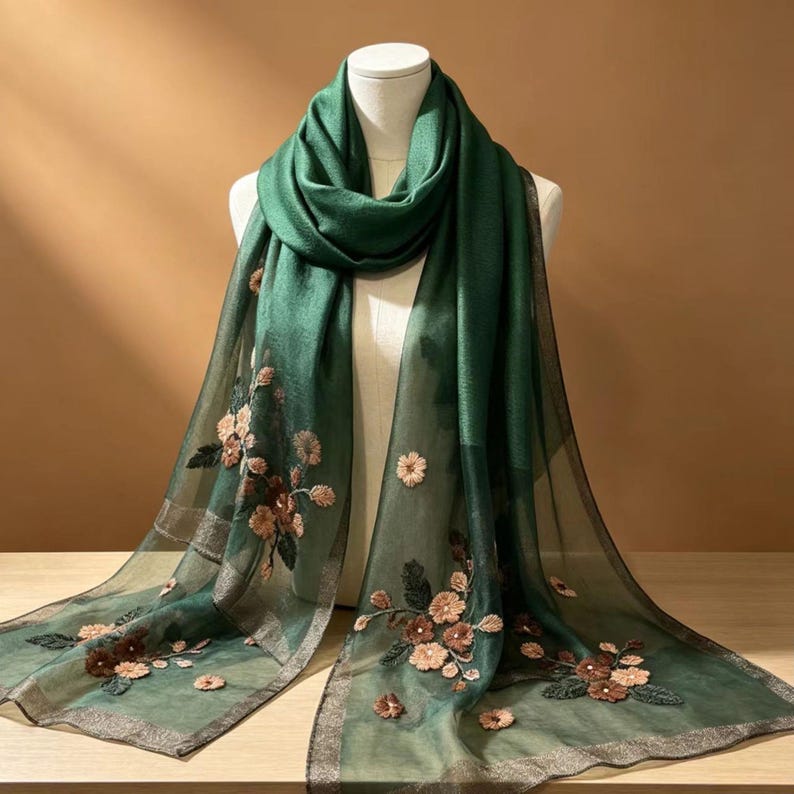 May include: A dark green scarf with embroidered floral details. The scarf is draped over a mannequin, showcasing its flowing design. The floral embroidery features shades of brown and pink, adding a delicate touch to the accessory.