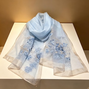 May include: A light blue scarf with embroidered blue floral designs and a gold trim. The scarf is made of a sheer, lightweight fabric. The scarf is draped over a white surface.