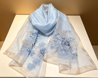 May include: A light blue scarf with embroidered blue floral designs and a gold trim. The scarf is made of a sheer, lightweight fabric. The scarf is draped over a white surface.