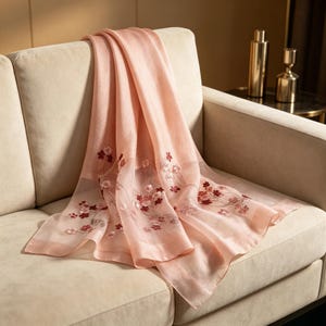 May include: A sheer, blush-pink scarf with embroidered floral details in shades of red and white. The scarf is draped over a beige sofa, showcasing its delicate, flowing fabric. The floral pattern is concentrated along the edges.