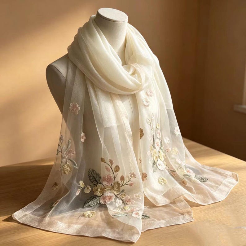 May include: A cream-colored, sheer scarf with embroidered floral details. The scarf is draped over a mannequin, showcasing the delicate embroidery of pink, yellow, and green flowers and leaves. The scarf's edges have a subtle, decorative trim.