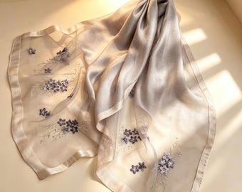 May include: A sheer, light gray scarf with embroidered purple floral accents. The scarf has a satin border and is draped to show its flowing texture. The delicate embroidery adds a touch of elegance to this accessory.