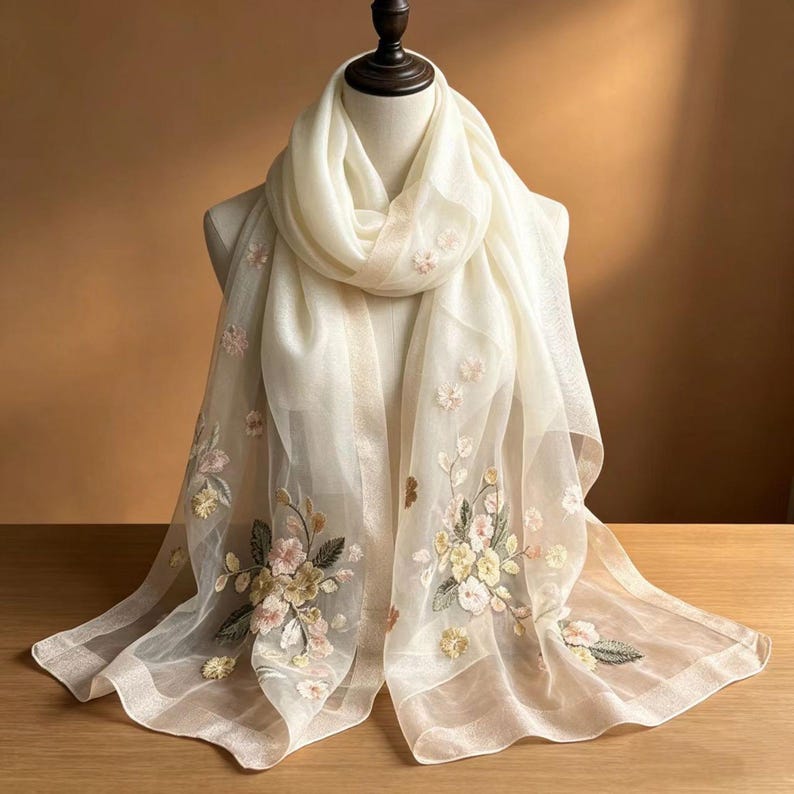 May include: A cream-colored scarf with floral embroidery. The scarf is draped over a mannequin, showcasing its delicate, sheer fabric. The embroidery features pink, yellow, and green flowers and leaves, adding a touch of elegance. The scarf's edges are finished with a subtle trim.