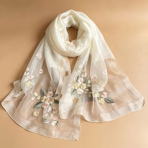 May include: A cream-colored, sheer scarf with embroidered floral designs in shades of pink, yellow, and green. The scarf is draped, showcasing the delicate fabric and detailed embroidery. The scarf is a fashion accessory.