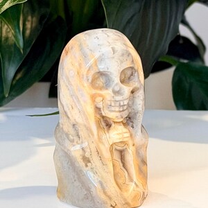Pode incluir: Uma escultura de cr&acirc;nio em pedra esculpida com um capuz drapeado. O cr&acirc;nio &eacute; detalhado com &oacute;rbitas oculares, um nariz e dentes. A pedra tem uma mistura de cores bege, cinza e castanho. A escultura &eacute; um item decorativo.