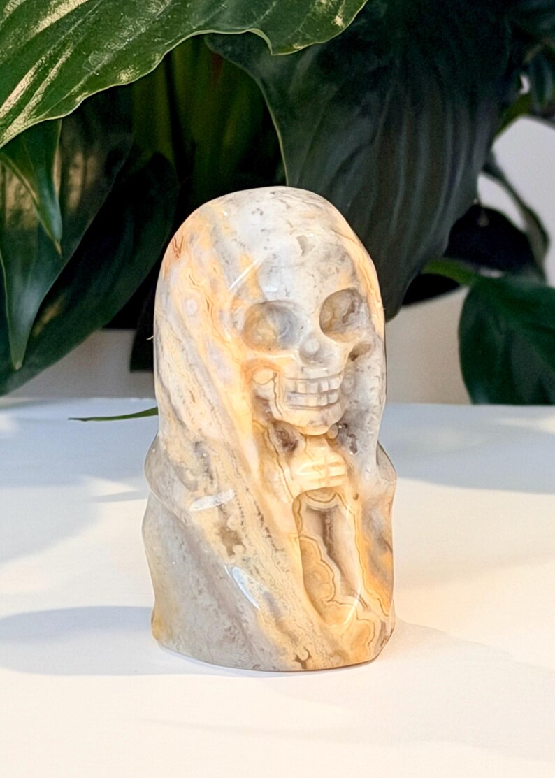 Pode incluir: Uma escultura de cr&acirc;nio em pedra esculpida com um capuz drapeado. O cr&acirc;nio &eacute; detalhado com &oacute;rbitas oculares, um nariz e dentes. A pedra tem uma mistura de cores bege, cinza e castanho. A escultura &eacute; um item decorativo.