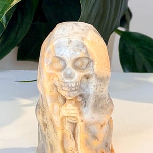 Pode incluir: Uma escultura de cr&acirc;nio em pedra esculpida com um capuz drapeado. O cr&acirc;nio &eacute; detalhado com &oacute;rbitas oculares, nariz e dentes. A pedra tem uma mistura de tons bege, cinza e laranja. A escultura &eacute; um item decorativo.