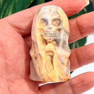 Pode incluir: Uma estatueta de caveira de pedra esculpida com uma capa com capuz. A caveira tem &oacute;rbitas oculares, uma cavidade nasal e dentes. A pedra tem uma mistura de cores bege, creme e cinza, com um padr&atilde;o em espiral. A estatueta &eacute; segurada em uma m&atilde;o.