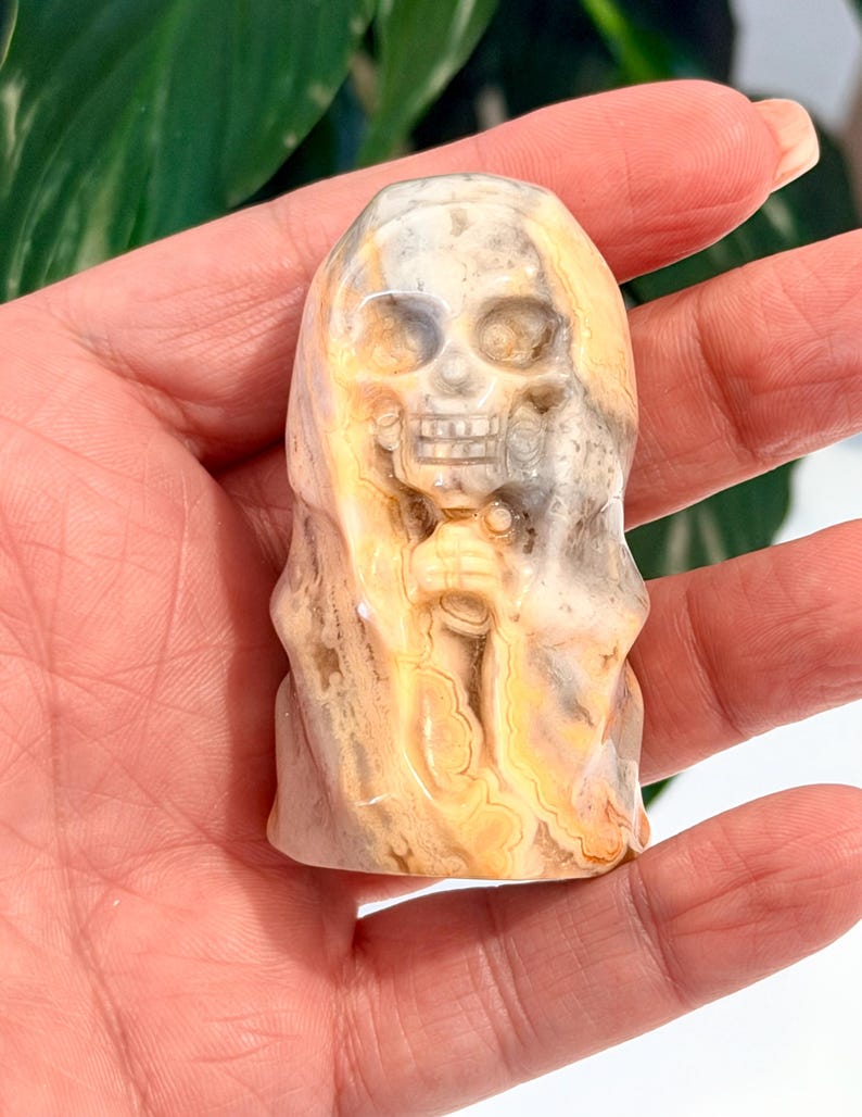 Pode incluir: Uma estatueta de caveira de pedra esculpida com uma capa com capuz. A caveira tem &oacute;rbitas oculares, uma cavidade nasal e dentes. A pedra tem uma mistura de cores bege, creme e cinza, com um padr&atilde;o em espiral. A estatueta &eacute; segurada em uma m&atilde;o.