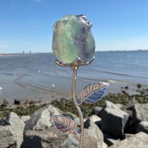 May include: A decorative rose sculpture with a green and purple fluorite rosebud and silver-colored stem and leaves. The sculpture is set against a backdrop of a beach and blue sky.