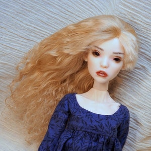May include: A doll with long, curly, blonde hair wearing a blue dress with a floral pattern. The doll has a porcelain face with delicate features and closed eyes.