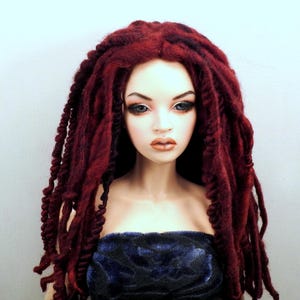May include: A doll with a pale complexion, blue eyes, and full lips. The doll has long, dark red dreadlocks and is wearing a dark blue strapless top. The background is a plain white.