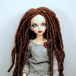 May include: A doll with a pale complexion, blue eyes, and dark red lips. The doll has long, brown dreadlocks and wears a gray shirt with a floral pattern and a red flower accent.