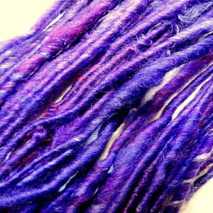 May include: A close-up of a bundle of purple and white synthetic dreadlocks. The dreadlocks are made of a soft, textured material and have a slightly wavy appearance.