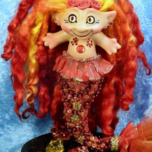 May include: A whimsical mermaid doll with vibrant red and yellow hair. The doll has a smiling face, a red bodice adorned with gems, and a beaded tail. It is decorated with sparkling embellishments and rests on a black base with colorful gems.