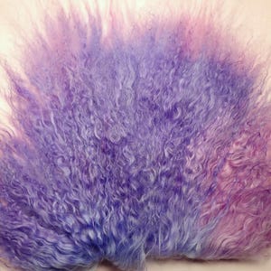 May include: A close-up of a fluffy, textured material in shades of purple and pink. The material has a wavy, curly appearance, with long, flowing strands. The colors blend together, creating a soft, ethereal look. The background is a light beige color.